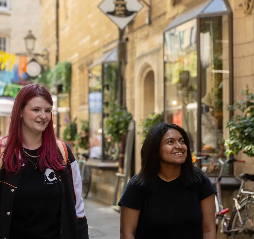Two University of Cambridge students are walking through Rose Crescent, central Cambridge, looking at shop windows while talking and smiling. Copyright: University of Cambridge (June 2024)