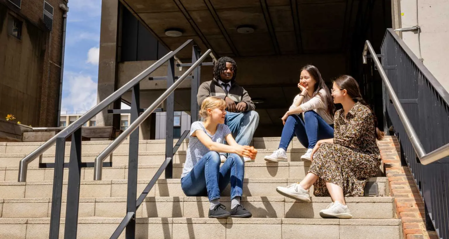 A group of happy University of Cambridge students sitting on stairs and chatting in the sunshine in front of the Museum of Zoology. Copyright: University of Cambridge (June 2024)