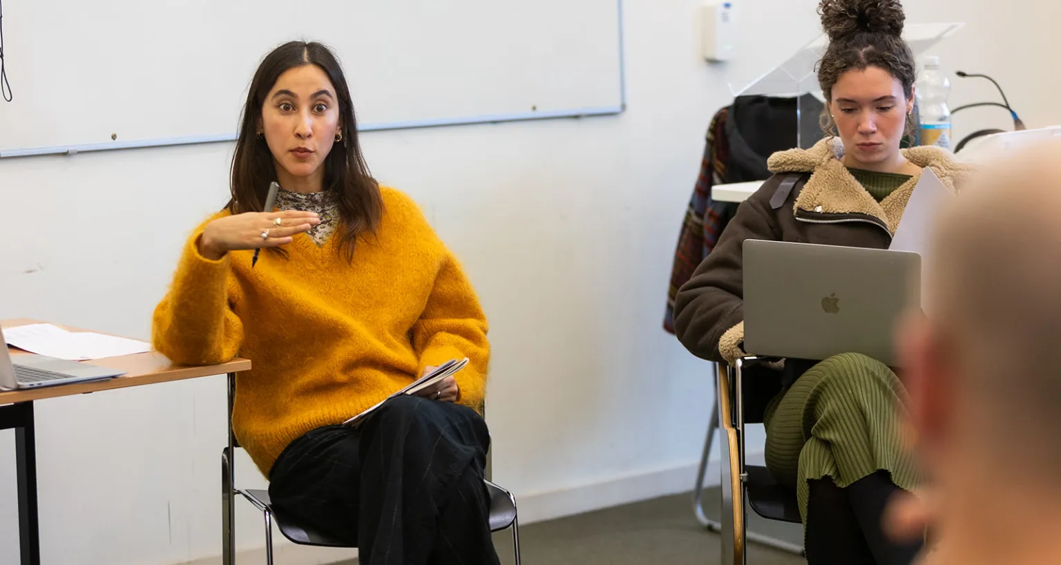 Lecturer sitting down, gesturing, wearing yellow jumper