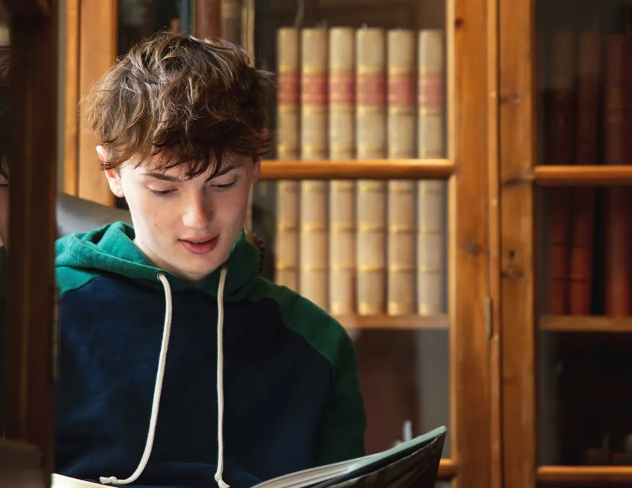 A University of Cambridge student reading a book in front of a bookcase at the Faculty of Architecture and History of Art library. Copyright: University of Cambridge (June 2023)