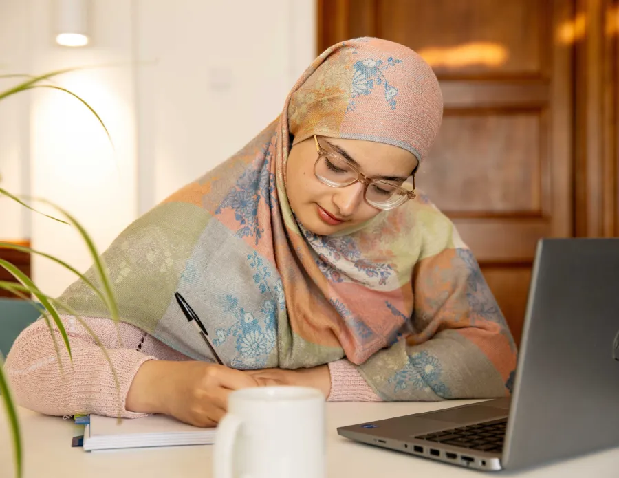 A University of Cambridge student writing in a notebook beside a laptop at the Student Services Centre (SSC). Copyright: University of Cambridge (June 2025).
