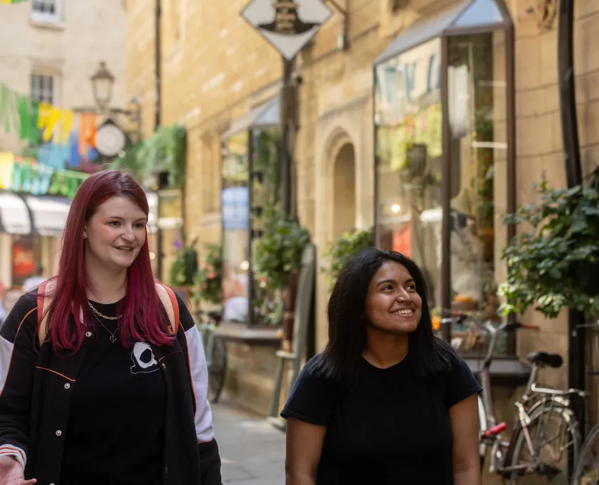 Two University of Cambridge students are walking through Rose Crescent, central Cambridge, looking at shop windows while talking and smiling. Copyright: University of Cambridge (June 2024)